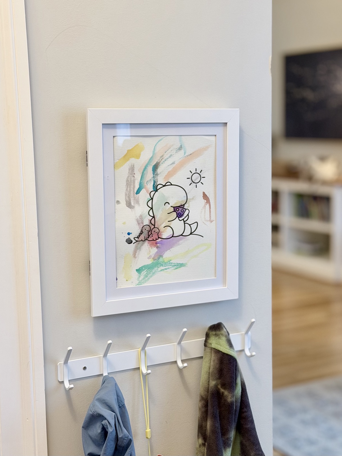A child's painted dinosaur drawing displayed in a white magnetic frame above coat hooks